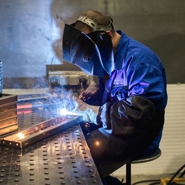 A welder in a blue uniform and protective mask works intently on metal parts, with bright sparks flying. The workshop has a focused, industrious atmosphere.
