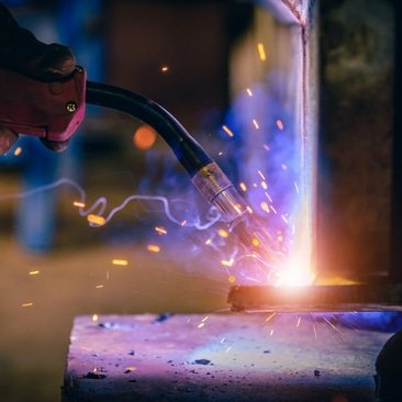 A welder works on steel fabrication, producing bright sparks during the welding process.